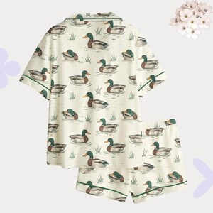 May include: Cream-colored pajama set featuring a pattern of ducks swimming in water. The short-sleeved top and shorts have green trim. The ducks are in shades of brown, green, and white.