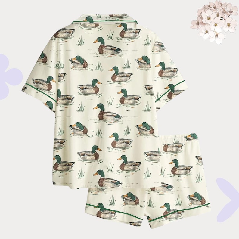 May include: Cream-colored pajama set featuring a pattern of ducks swimming in water. The short-sleeved top and shorts have green trim. The ducks are in shades of brown, green, and white.