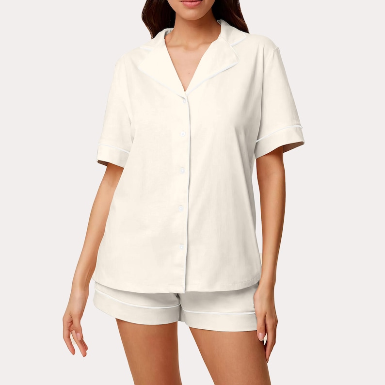 以下が含まれることがあります： クリーム色のパジャマセット。半袖のボタンダウンシャツと、お揃いのショートパンツが特徴です。シャツ、襟、袖、ショートパンツの縁には白いパイピングが施されています。生地は柔らかく、軽量な素材のようです。
