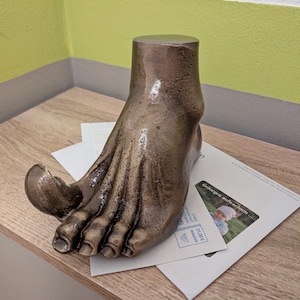 May include: A bronze-colored sculpture of a human foot, with detailed toes and a smooth, polished finish. The foot is resting on a wooden surface, with a white envelope and a small photo visible.