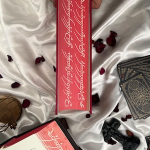 Puede incluir: Un libro rojo con texto blanco en el lomo, sobre una superficie de sat&eacute;n blanco salpicada de p&eacute;talos de rosa. El lomo del libro presenta una inscripci&oacute;n en una fuente estilizada. Tambi&eacute;n hay cartas del tarot y una cruz decorativa.