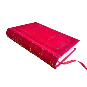 Op de afbeelding: Een rood hardcover boek met gouden letters en een bijpassend rood lint als bladwijzer. De kaft van het boek toont de titel en de naam van de auteur in goud. De rug heeft gouden banden en de naam van de auteur.