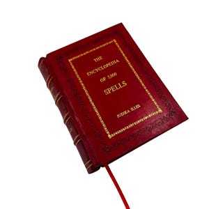 May include: A red leather-bound book titled "The Encyclopedia of 5,000 Spells" with gold lettering and decorative borders. A red ribbon bookmark extends from the book's spine. The author's name, Judika Illes, is also printed on the cover.