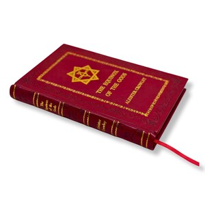 Op de afbeelding: Een rood hardcover boek getiteld "The Equinox of the Gods" van Aleister Crowley. De kaft heeft gouden letters en een stersymbool. Een rood lint-bladwijzer steekt uit het boek.