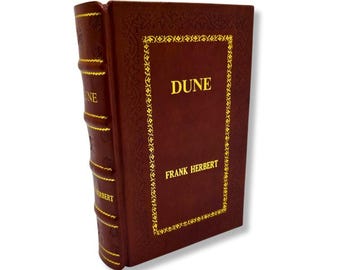 Puede incluir: Un libro de tapa dura titulado "DUNE" de Frank Herbert. El libro tiene una cubierta marrón oscuro con letras doradas y un borde decorativo. La columna vertebral también presenta el título y el nombre del autor en oro.