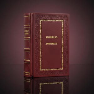 Puede incluir: Un libro de color burdeos con letras y adornos dorados. La portada dice "Alcoholics Anonymous" en oro. El lomo también tiene letras doradas. El libro está de pie y se refleja en la superficie de abajo.