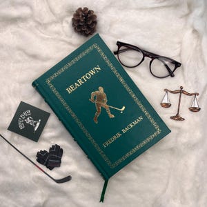 May include: A green hardcover book titled "Beartown" with gold lettering and a hockey player illustration. The image includes a pair of glasses, a hockey stick, gloves, and a scale of justice pin.