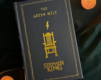 Pode incluir: Um livro de capa dura cinza escuro intitulado "The Green Mile" de Stephen King. O livro tem letras douradas e um gráfico de cadeira elétrica dourada. A capa é emoldurada com uma borda dourada. Duas velas acesas no fundo.