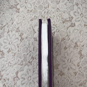 May include: A hardcover book with a purple cover and silver-edged pages. The spine is a deep purple, and the pages are a bright white. The book is set against a white lace background.