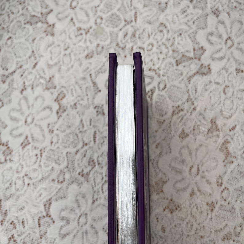May include: A hardcover book with a purple cover and silver-edged pages. The spine is a deep purple, and the pages are a bright white. The book is set against a white lace background.