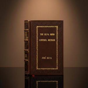 Puede incluir: Un libro de tapa dura titulado "THE SILVA MIND CONTROL METHOD" de José Silva. El libro tiene una cubierta marrón con letras doradas y bordes decorativos. El lomo también tiene letras doradas. El libro se refleja en una superficie oscura.