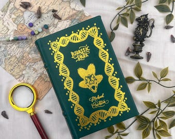 Puede incluir: Un libro verde titulado "Jurassic Park" con letras doradas y un diseño de ADN. Una lupa, un mapa y elementos decorativos rodean el libro. El nombre del autor, Michael Crichton, también está en la portada.