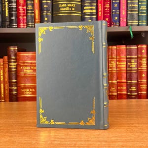K&ouml;nnte beinhalten: Ein blaugr&uuml;nes Buch mit goldenen dekorativen Eckdesigns und goldenen Verzierungen. Das Buch steht auf einer Holzoberfl&auml;che, mit einem B&uuml;cherregal im Hintergrund. Der Buchr&uuml;cken ist sichtbar und die Seiten sind geschlossen.