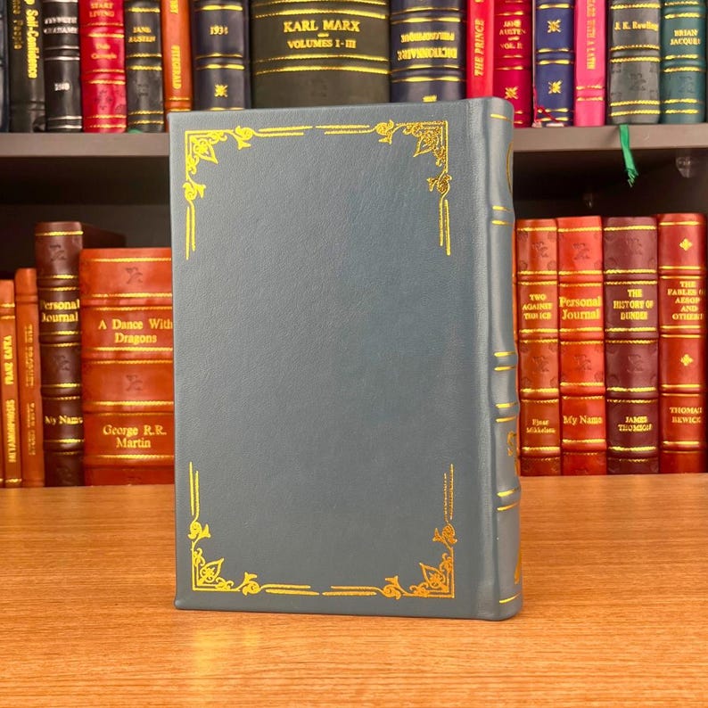 K&ouml;nnte beinhalten: Ein blaugr&uuml;nes Buch mit goldenen dekorativen Eckdesigns und goldenen Verzierungen. Das Buch steht auf einer Holzoberfl&auml;che, mit einem B&uuml;cherregal im Hintergrund. Der Buchr&uuml;cken ist sichtbar und die Seiten sind geschlossen.