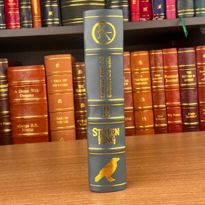 K&ouml;nnte beinhalten: Buchr&uuml;cken von "The Drawing of the Three" von Stephen King. Der R&uuml;cken ist grau mit goldener Schrift und einer goldenen Rabenillustration. Weitere B&uuml;cher sind im Hintergrund zu sehen.