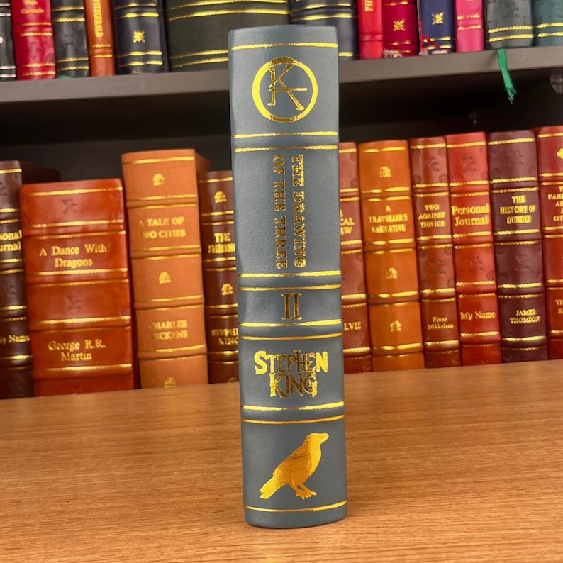 K&ouml;nnte beinhalten: Buchr&uuml;cken von "The Drawing of the Three" von Stephen King. Der R&uuml;cken ist grau mit goldener Schrift und einer goldenen Rabenillustration. Weitere B&uuml;cher sind im Hintergrund zu sehen.