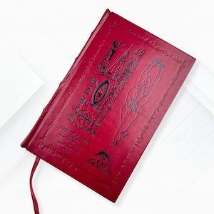 Puede incluir: Un libro rojo encuadernado en cuero titulado "Book of Pleasure" con jerogl&iacute;ficos e ilustraciones en relieve. La portada presenta un s&iacute;mbolo de ojo y una figura. Una cinta roja de marcador es visible.