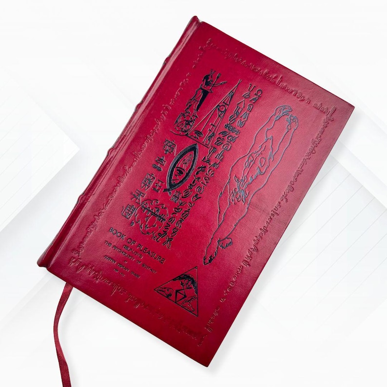 Puede incluir: Un libro rojo encuadernado en cuero titulado "Book of Pleasure" con jerogl&iacute;ficos e ilustraciones en relieve. La portada presenta un s&iacute;mbolo de ojo y una figura. Una cinta roja de marcador es visible.