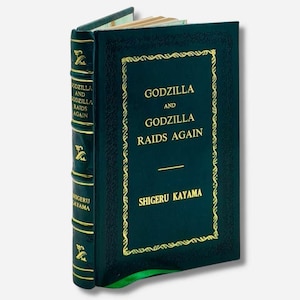 Può includere: Un libro con copertina rigida intitolato "Godzilla and Godzilla Raids Again" di Shigeru Kayama. Il libro ha una copertina verde scuro con scritte dorate e un bordo decorativo. Il dorso presenta anche scritte e abbellimenti dorati.