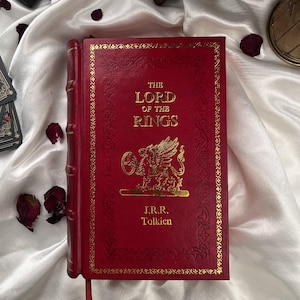 Puede incluir: Un libro de tapa dura rojo titulado "The Lord of the Rings" con letras doradas en relieve y una ilustraci&oacute;n detallada. El libro presenta un borde decorativo y el nombre del autor, J.R.R. Tolkien.