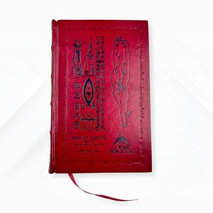 Puede incluir: Un libro rojo con ilustraciones y texto en relieve. La portada presenta s&iacute;mbolos, figuras y el t&iacute;tulo "BOOK OF PLEASURE". Una cinta roja de marcador se extiende desde el lomo del libro. Los bordes del libro tambi&eacute;n son rojos.