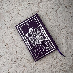 May include: A purple book with a silver graphic of a cat, spiderweb, and open windows. A purple ribbon is attached to the book. The book's cover has a gothic theme.