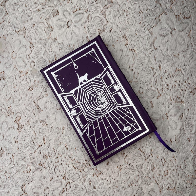 May include: A purple book with a silver graphic of a cat, spiderweb, and open windows. A purple ribbon is attached to the book. The book's cover has a gothic theme.