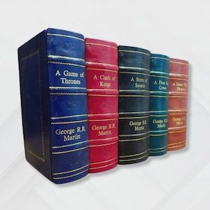May include: A collection of five hardcover books with gold lettering and accents. The spines are in various colors: blue, red, black, teal, and orange. The titles include "A Game of Thrones" and "A Clash of Kings" by George R.R. Martin.