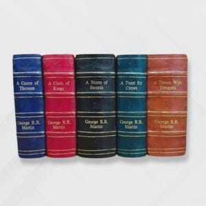 May include: A collection of five books with titles from the "A Song of Ice and Fire" series by George R.R. Martin. The books are bound in various colors: blue, red, black, teal, and brown. Each book spine displays the title and author's name.