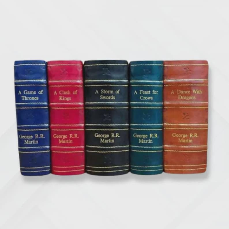 May include: A collection of five books with titles from the "A Song of Ice and Fire" series by George R.R. Martin. The books are bound in various colors: blue, red, black, teal, and brown. Each book spine displays the title and author's name.