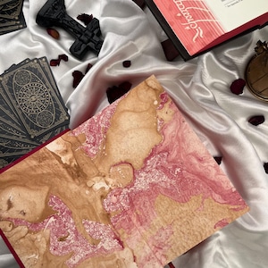 Puede incluir: Un libro abierto con p&aacute;ginas veteadas en rosa y beige, una baraja de cartas del tarot y una cruz decorativa negra descansan sobre una superficie de sat&eacute;n blanco. P&eacute;talos de rosa rojos dispersos a&ntilde;aden color.