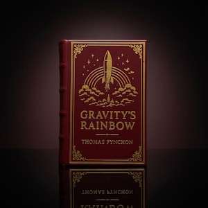 May include: A hardcover book titled "Gravity's Rainbow" by Thomas Pynchon. The cover is burgundy with gold foil accents, featuring a rocket launching over a rainbow. The book's title and author's name are in gold lettering.