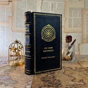 Peut inclure: Un livre à couverture rigide noire intitulé "His Dark Materials" de Philip Pullman, avec des lettres dorées en relief et un motif circulaire décoratif. La tranche comporte également des lettres dorées. Le livre est présenté sur une carte vintage.