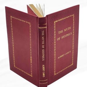May include: A burgundy hardcover book, open to display its pages. The cover features gold decorative borders and the title "THE MYTH OF SISYPHUS" with the author, ALBERT CAMUS, in gold lettering. The page edges are also gold.