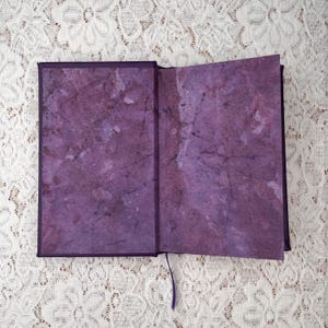 May include: An open journal with a purple cover and matching interior pages. The pages have a marbled texture in shades of purple and white. A purple ribbon bookmark is visible.