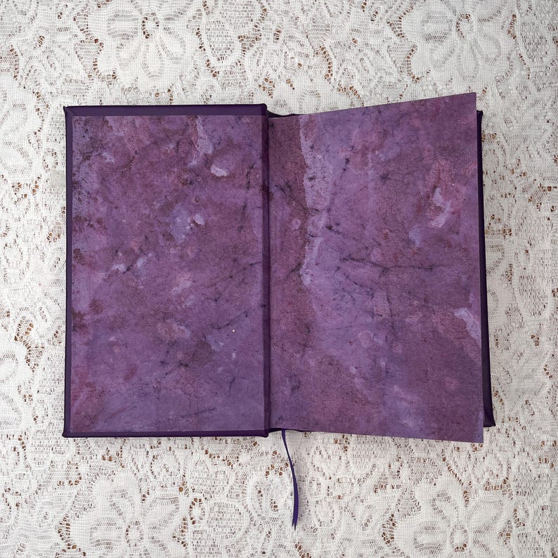 May include: An open journal with a purple cover and matching interior pages. The pages have a marbled texture in shades of purple and white. A purple ribbon bookmark is visible.