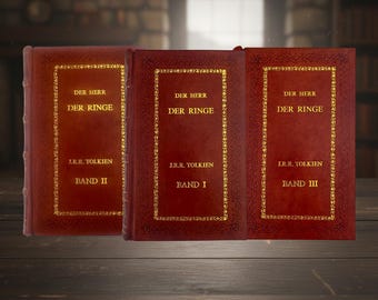 May include: Three hardback books with red covers and gold lettering. The titles "DER HERR DER RINGE" and "J.R.R. Tolkien" are visible. The books are labelled "BAND I", "BAND II", and "BAND III". The books are displayed on a wooden surface.