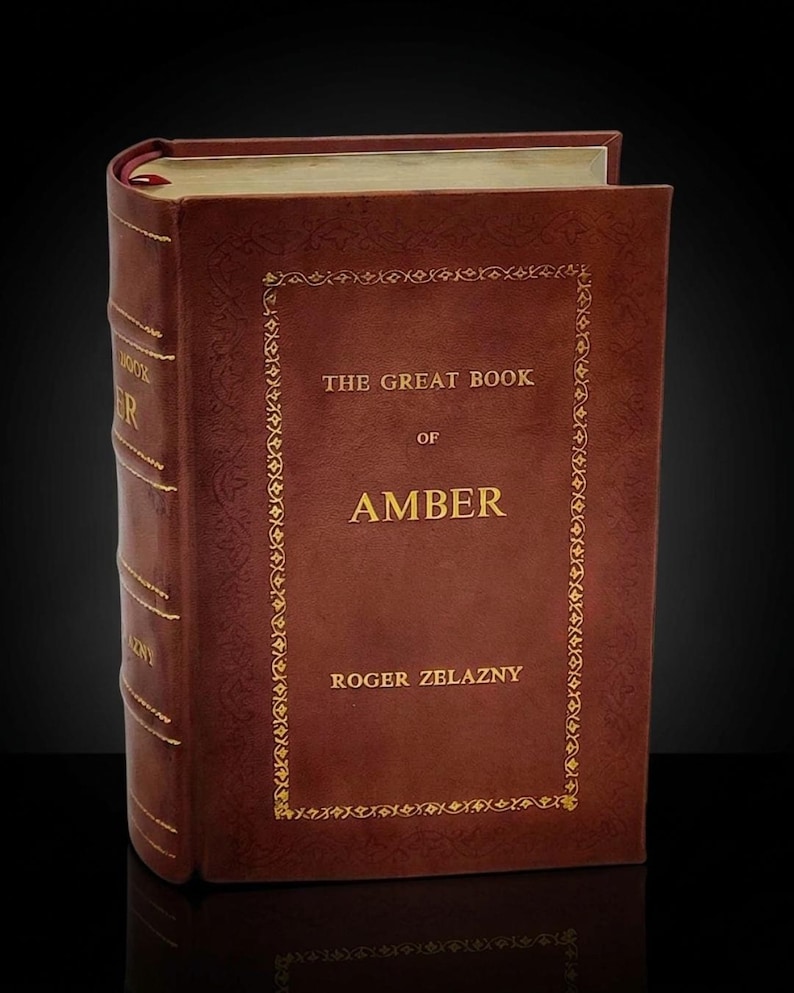 May include: A hardcover book titled "THE GREAT BOOK OF AMBER" by Roger Zelazny. The book has a brown leather cover with gold lettering and decorative border. A red ribbon bookmark is visible.