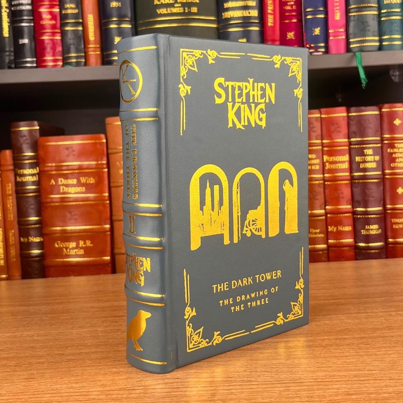 K&ouml;nnte beinhalten: Ein Hardcover-Buch mit dem Titel "The Dark Tower: The Drawing of the Three" von Stephen King. Das Buch hat einen grauen Einband mit goldenen Buchstaben und Illustrationen. Auf dem Buchr&uuml;cken ist eine Kr&auml;he abgebildet. Weitere B&uuml;cher sind im Hintergrund zu sehen.