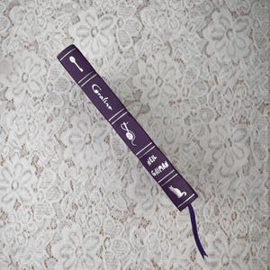 May include: A purple book titled "Coraline" by Neil Gaiman, with white text and illustrations of a key, a mouse, and a cat. A purple ribbon bookmark extends from the bottom of the book. The book is on a white lace surface.