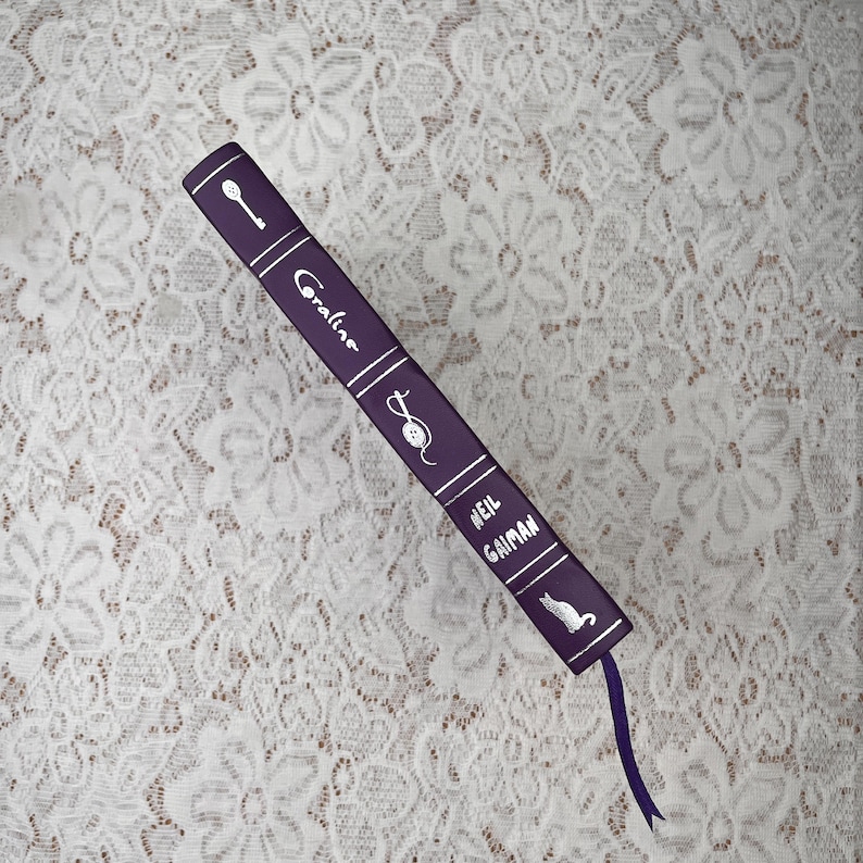 May include: A purple book titled "Coraline" by Neil Gaiman, with white text and illustrations of a key, a mouse, and a cat. A purple ribbon bookmark extends from the bottom of the book. The book is on a white lace surface.