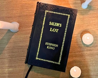 Pode incluir: Um livro de capa dura azul escuro intitulado "Salem's Lot" de Stephen King, com letras douradas e bordas decorativas. Uma fita preta como marcador de livros é visível. Várias velas acesas circundam o livro em uma superfície de madeira.