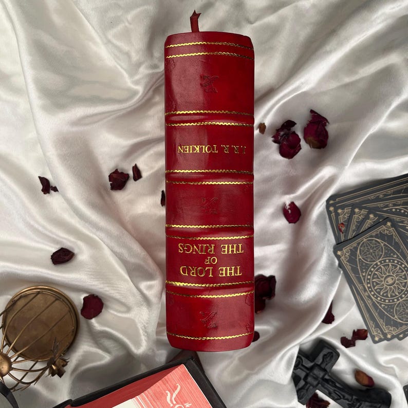 Puede incluir: Una vela roja con forma de libro, con letras doradas que dicen "THE LORD OF THE RINGS" y "J.R.R. TOLKIEN". La vela tiene bandas doradas y una mecha. P&eacute;talos de rosa secos y naipes en el fondo.