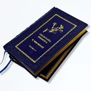 May include: A stack of dark blue hardback books with gold lettering and detailing. The covers display the title "Collectanea Hermetica" and the author's name, W. Wynn Westcott. A blue ribbon bookmark is visible.