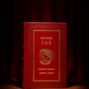 Op de afbeelding: Een rood hardcover boek met de titel "SAVAGE FAE" met gouden letters en een decoratieve rand. Het boek bevat een illustratie van twee gezichten en de namen van de auteurs, Caroline Peckham en Susanne Valenti. Een rood lint-bladwijzer is zichtbaar.