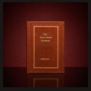 Op de afbeelding: Een hardcover boek getiteld "The Three-Body Problem" van Cixin Liu. Het boek heeft een bruine leren kaft met goudkleurige reliëftekst en een decoratieve rand. De achtergrond is donkerrood.