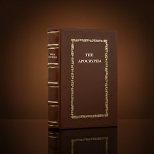 May include: A hardcover book titled "THE APOCRYPHA" with a brown leather cover and gold-colored accents. The spine features the text "THE OCRYP". The book is displayed on a reflective surface.