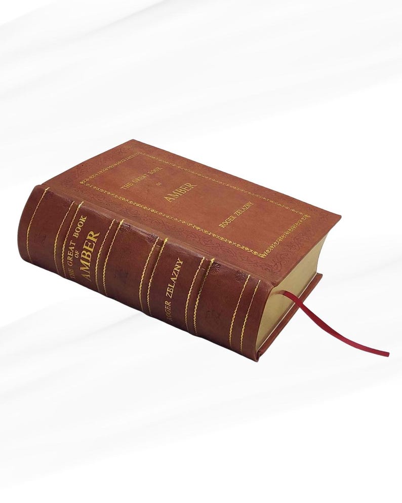 May include: A hardcover book titled "The Great Book of Amber" by Roger Zelazny. The book has a brown leather cover with gold lettering and detailing. A red ribbon bookmark is visible.