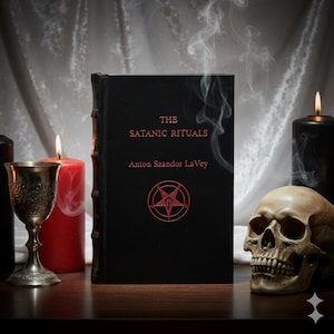 May include: A black book titled "The Satanic Rituals" by Anton Szandor LaVey, featuring a red pentagram symbol. The scene includes a skull, candles, and a chalice, creating a gothic aesthetic.