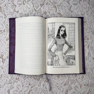 May include: An open book with a purple cover and a ribbon bookmark. The right page features a black and white illustration of a woman with large black eyes, wearing a long-sleeved top and apron. The text reads, "Her eyes were big black buttons."